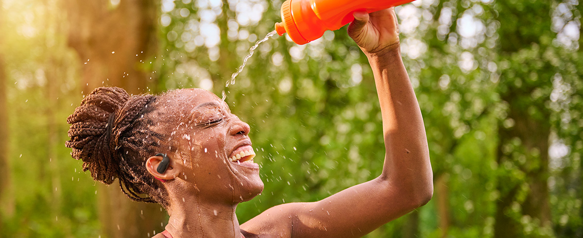 Frau k&uuml;hlt sich ab, indem sie sich Wasser &uuml;ber den Kopf gie&szlig;t, w&auml;hrend sie Philips Sportkopfh&ouml;rer im Freien tr&auml;gt